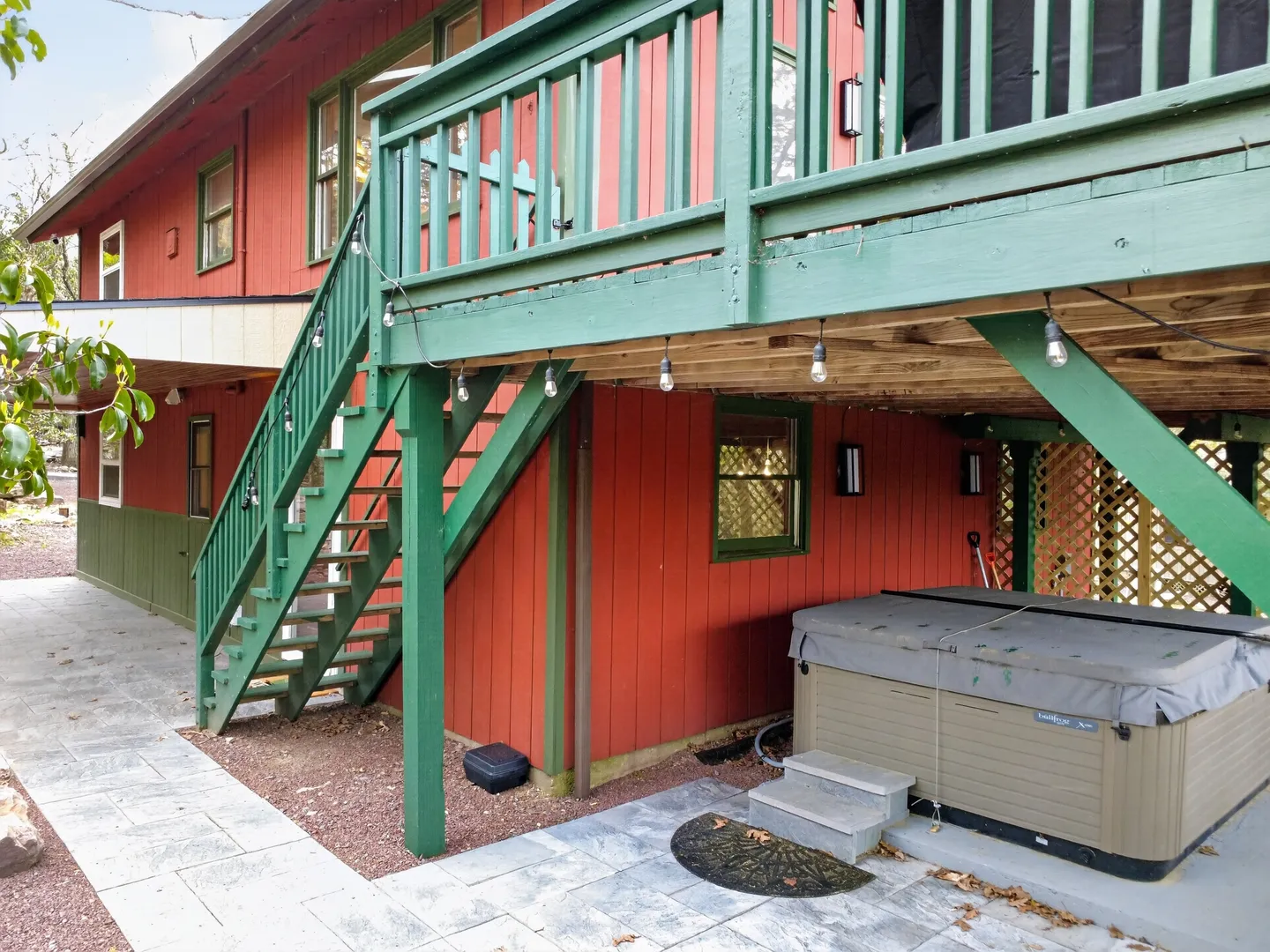 Exterior view of a red wood-sided house with a green wooden deck, stairs, and a covered hot tub on a stone patio.