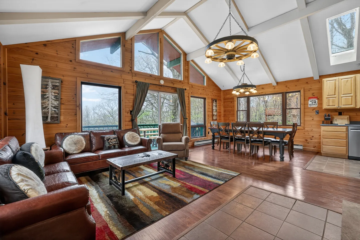 Open-concept living room and dining area featuring wood-paneled walls, vaulted ceiling with exposed beams, leather couches, wagon wheel chandeliers, and large windows.
