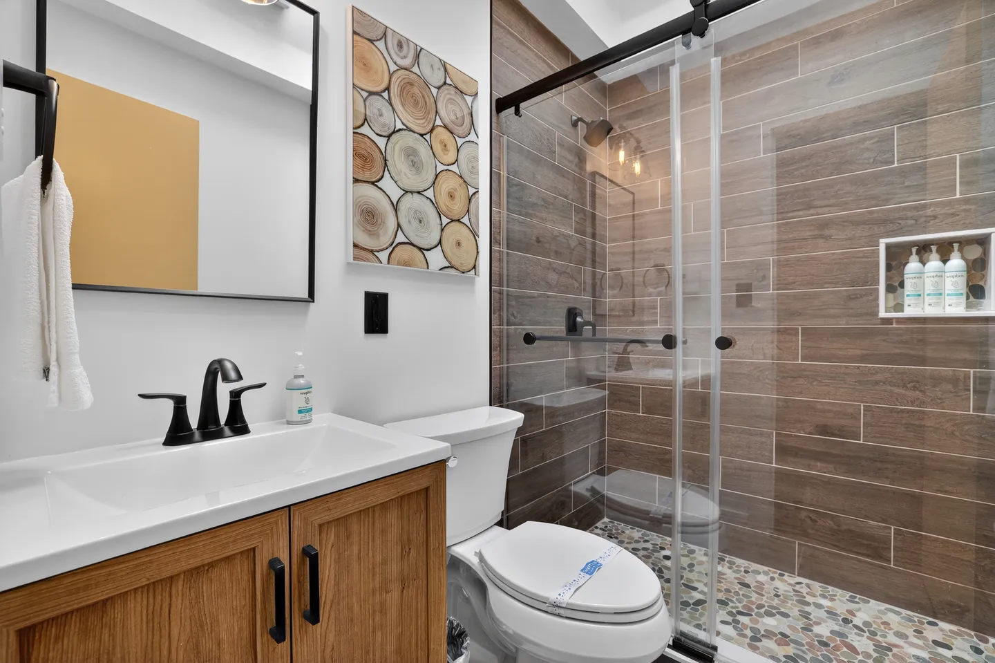 Bathroom with a wood-grain vanity, black fixtures, toilet, and a glass-enclosed walk-in shower with wood-look tile.