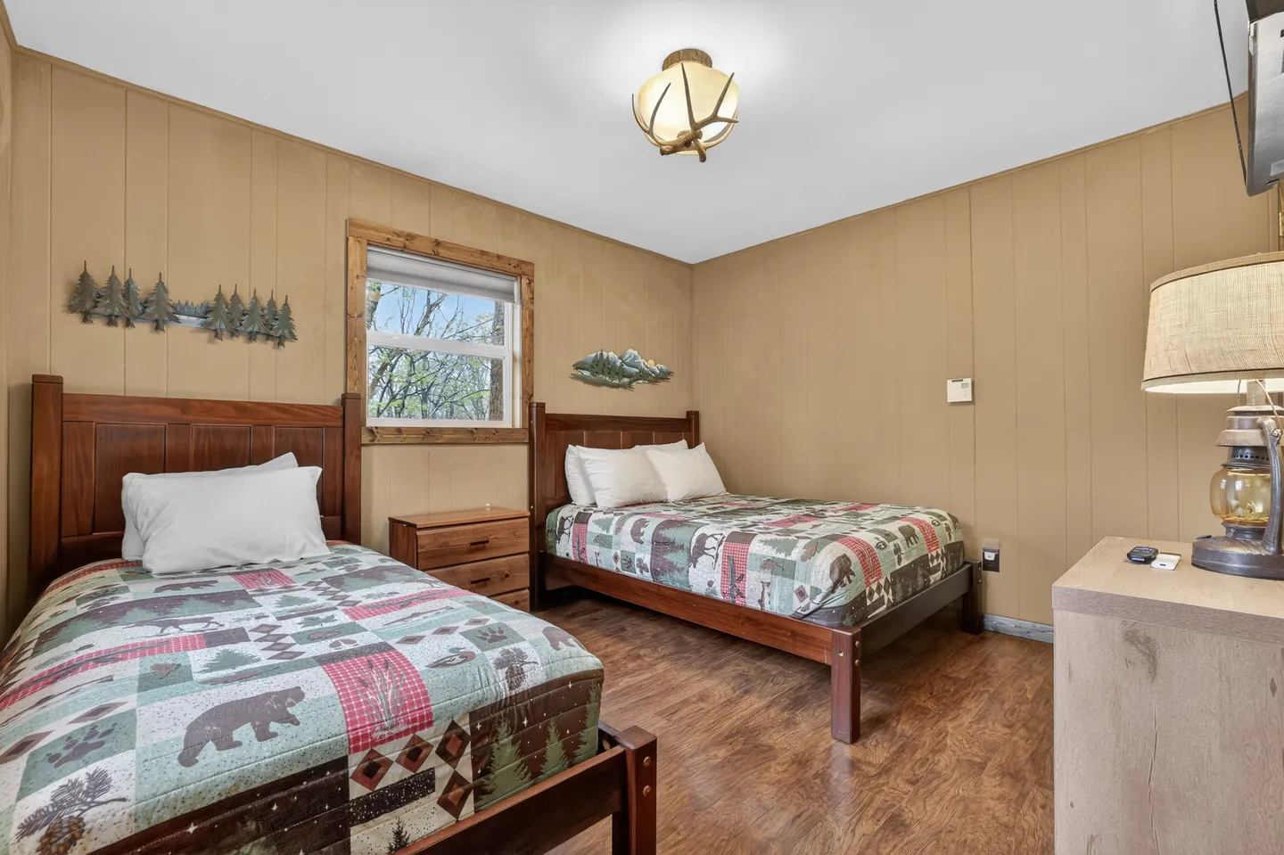 Bedroom with two beds topped with rustic wildlife-patterned quilts, wood-paneled walls, and an antler ceiling light.