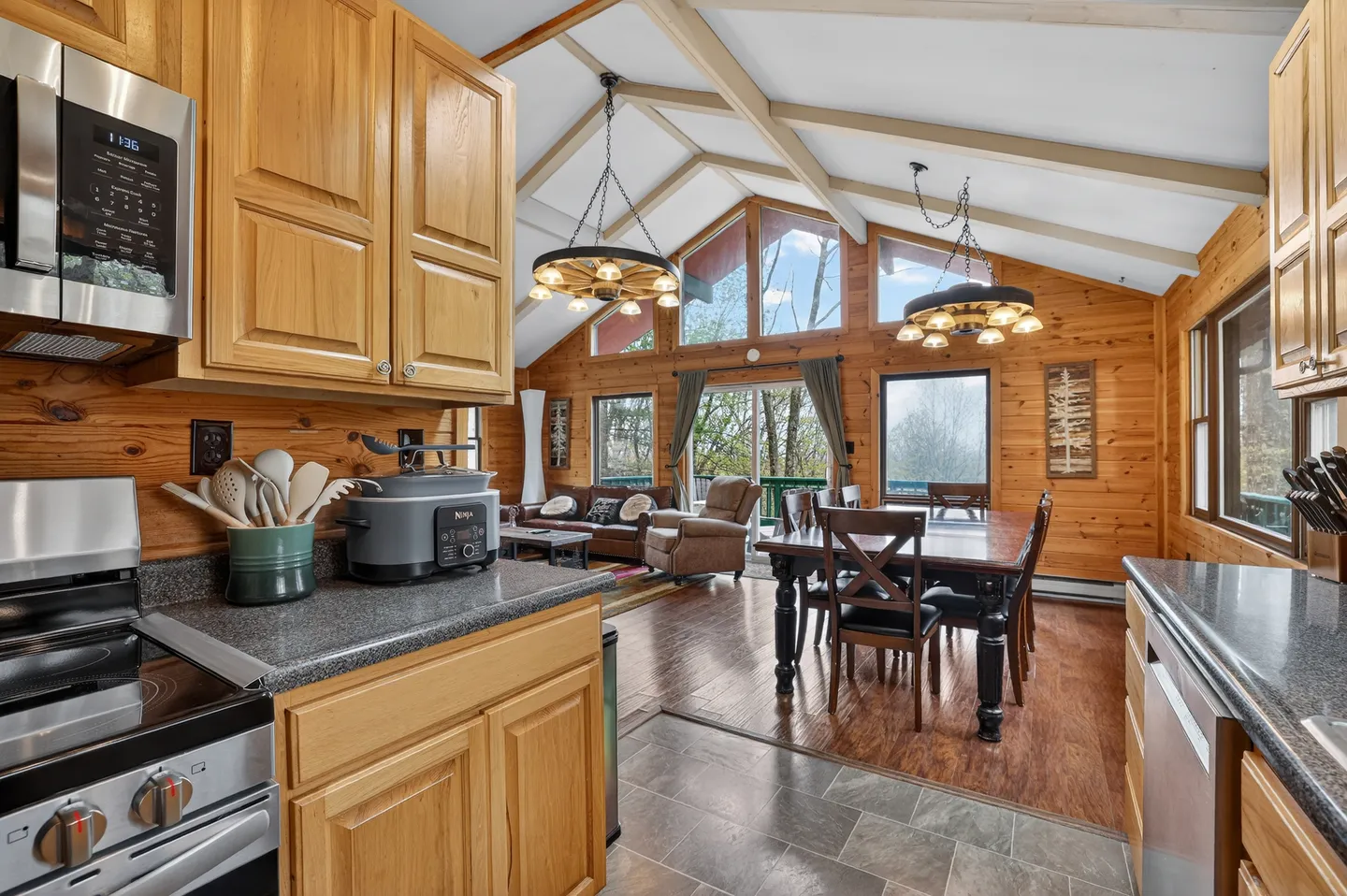 Open-concept cabin interior looking from the kitchen into the dining area and living room with vaulted ceilings and large windows.