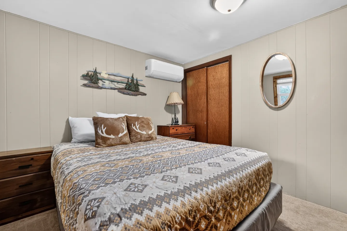 Bedroom with patterned quilt, antler-motif pillows, wall-mounted air conditioning unit, wood paneled closet doors, and an oval mirror.