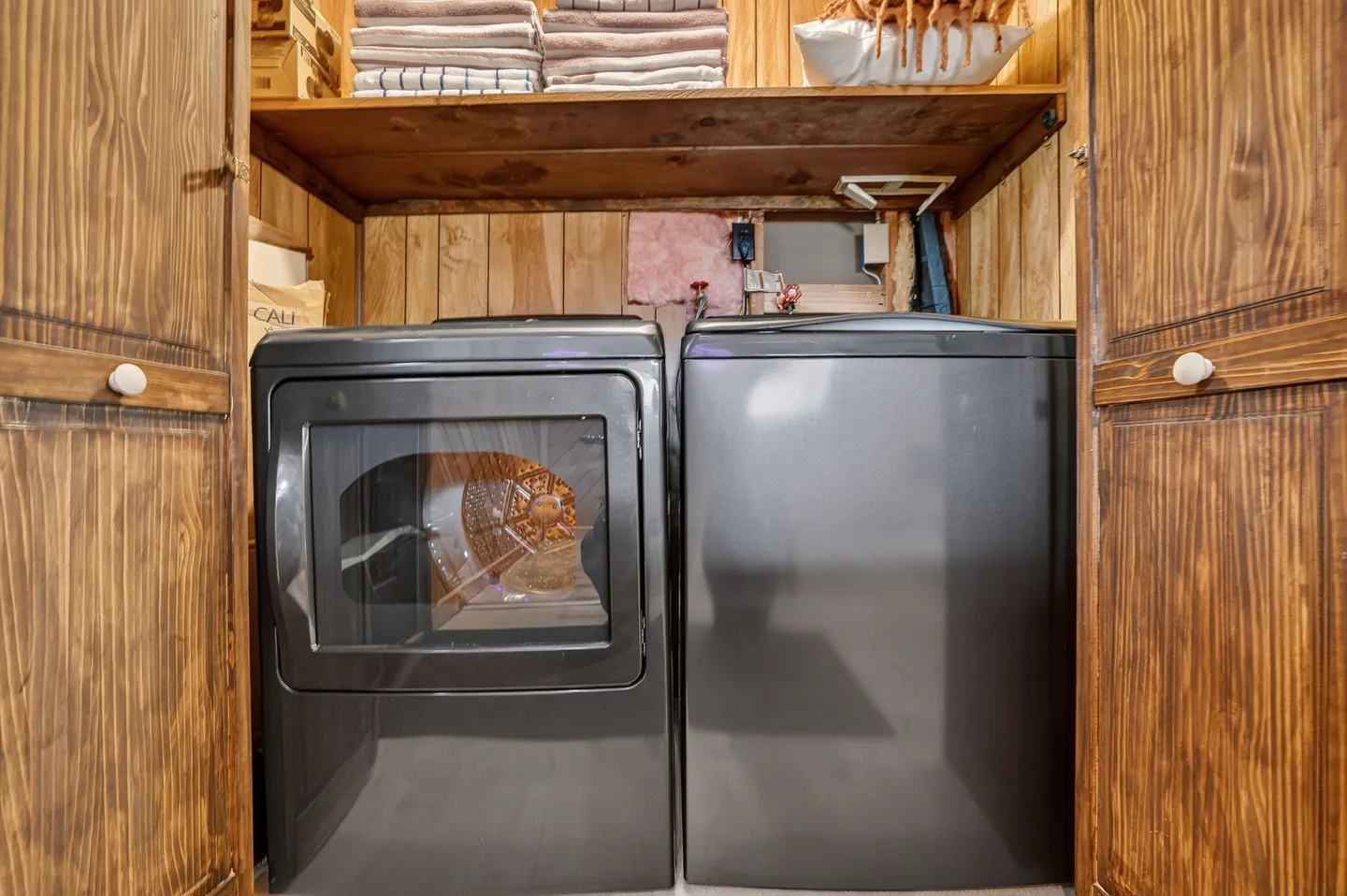A laundry closet with double wood doors containing a dark gray front-loading dryer, a top-loading washer, and an overhead shelf with folded linens.