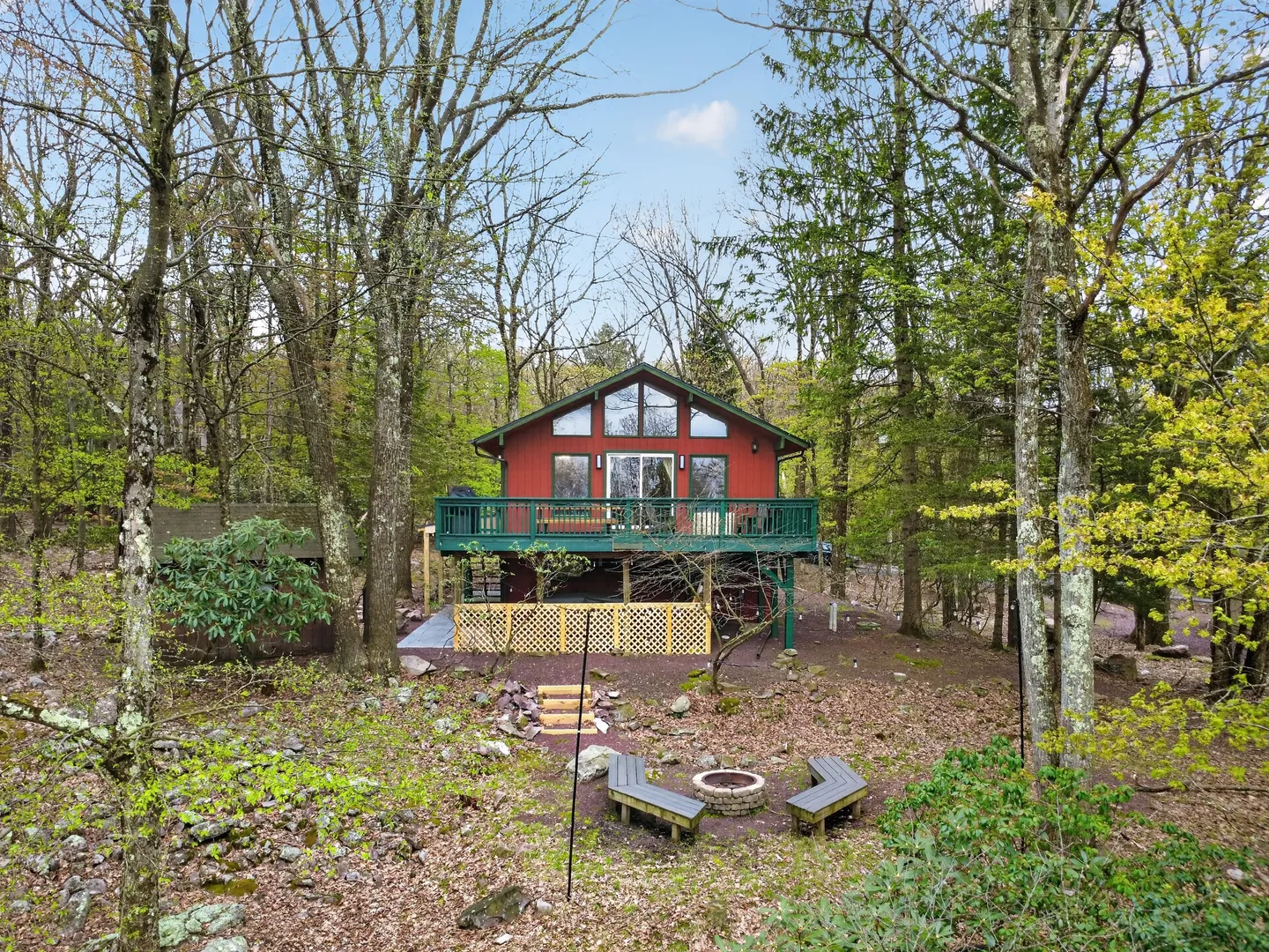 Exterior view of a red two-story cabin nestled in the woods with a green elevated deck and a fire pit in the yard.