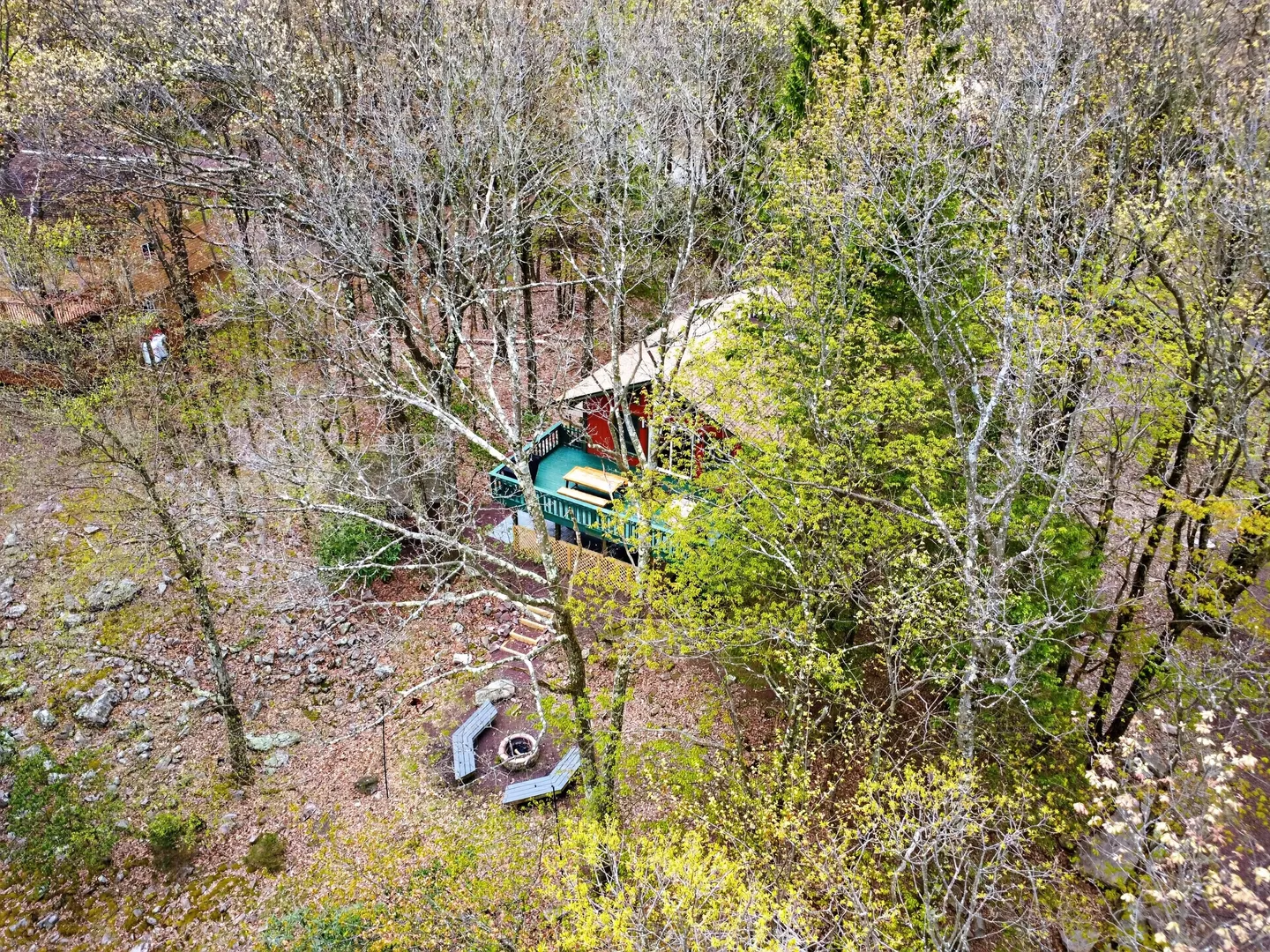 Aerial view looking down through trees at a cabin with a deck and an outdoor fire pit with benches.