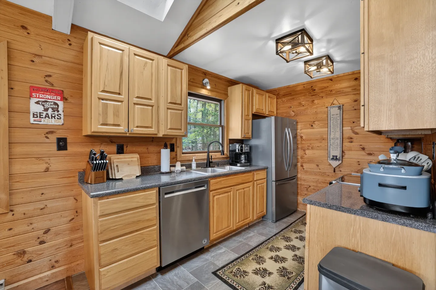 Kitchen with wood-paneled walls, light wood cabinets, stainless steel appliances, dark countertops, and a vaulted ceiling.