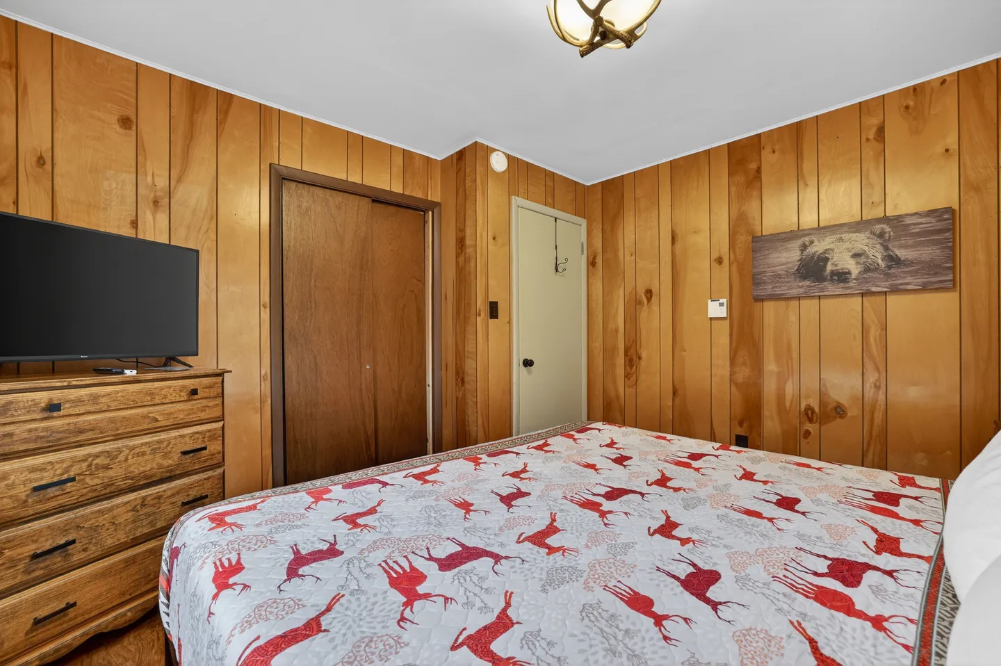 Bedroom with wood-paneled walls, a bed featuring a red and white deer-patterned bedspread, a wooden dresser with a flat-screen TV, and bear artwork on the wall.