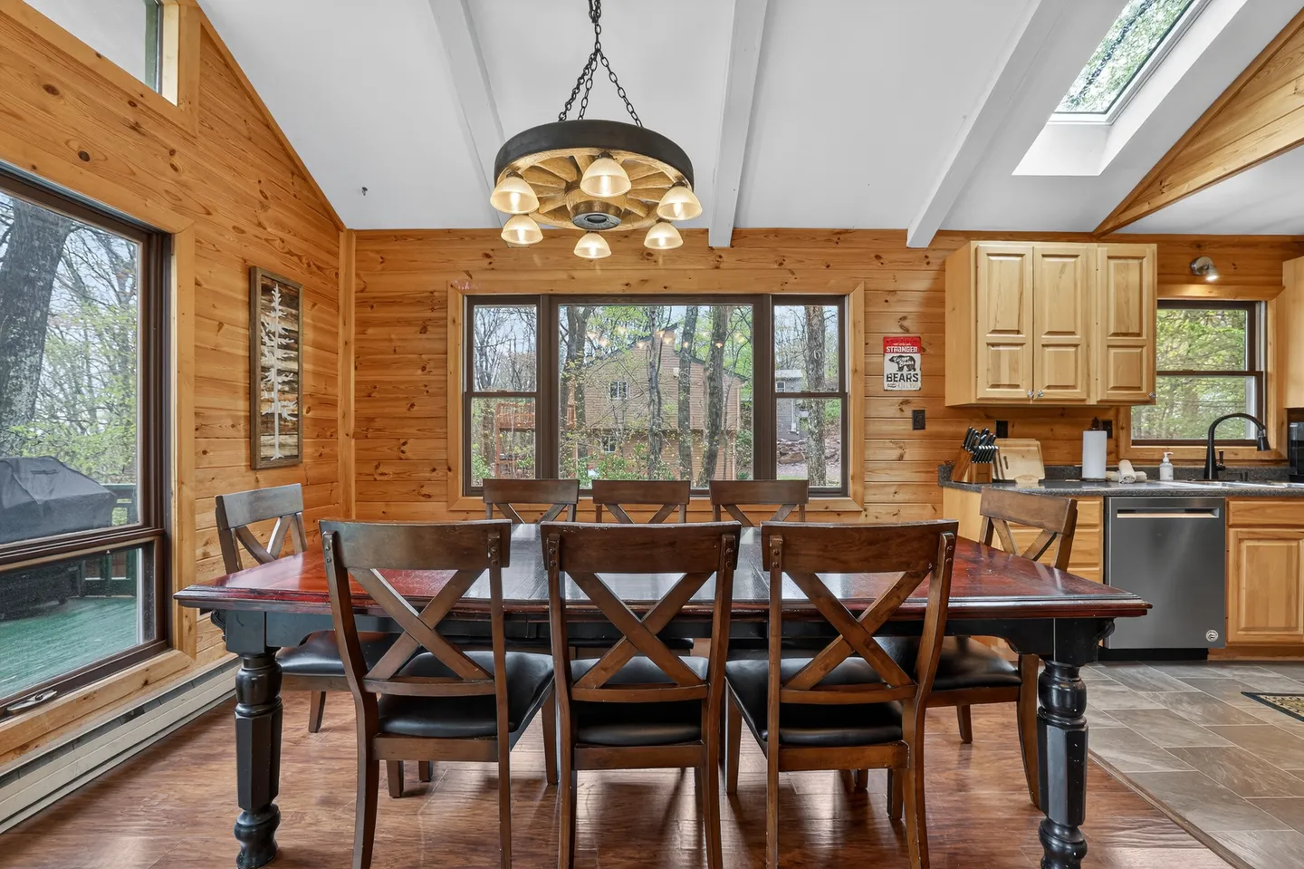 Dining area with a large wooden table, chairs, wagon-wheel chandelier, wood-paneled walls, and adjacent kitchen cabinets.