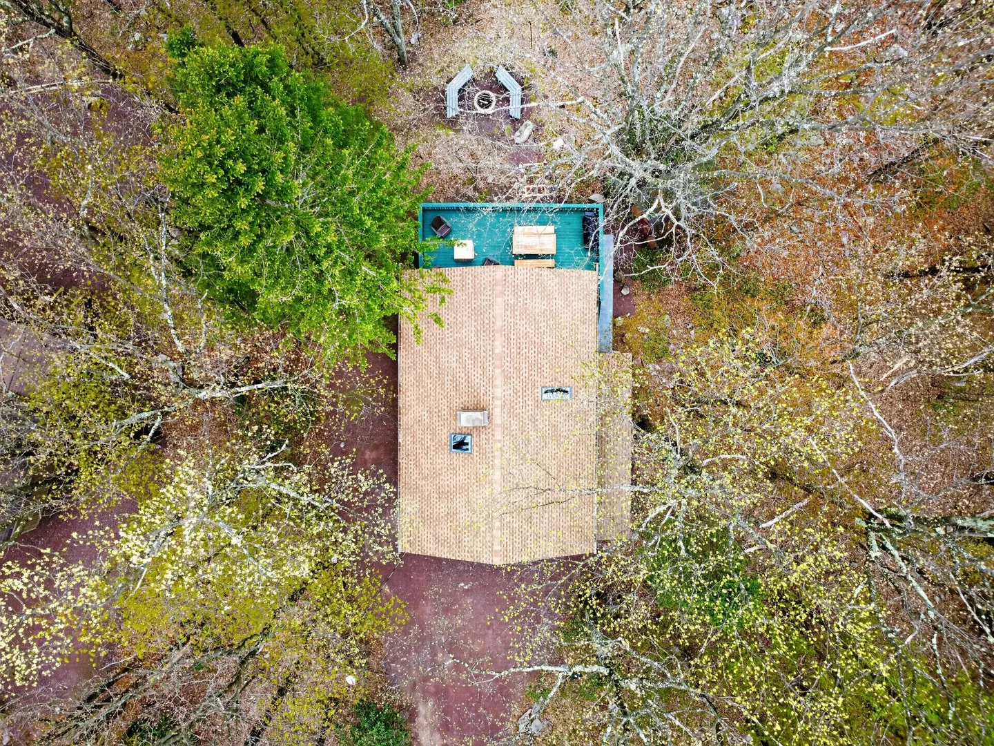 Overhead aerial view of a cabin roof, back deck, and fire pit nestled within a dense forest.