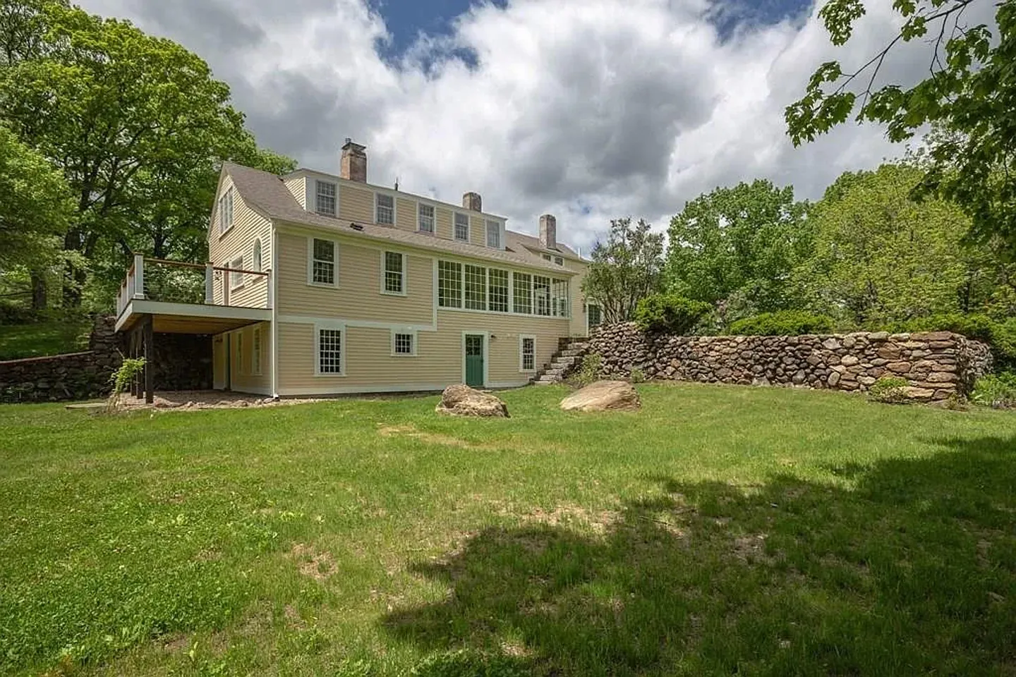 Rear exterior view of a multi-story yellow house with a green door, upper-level deck, stone retaining wall, and grassy lawn under a partly cloudy sky.