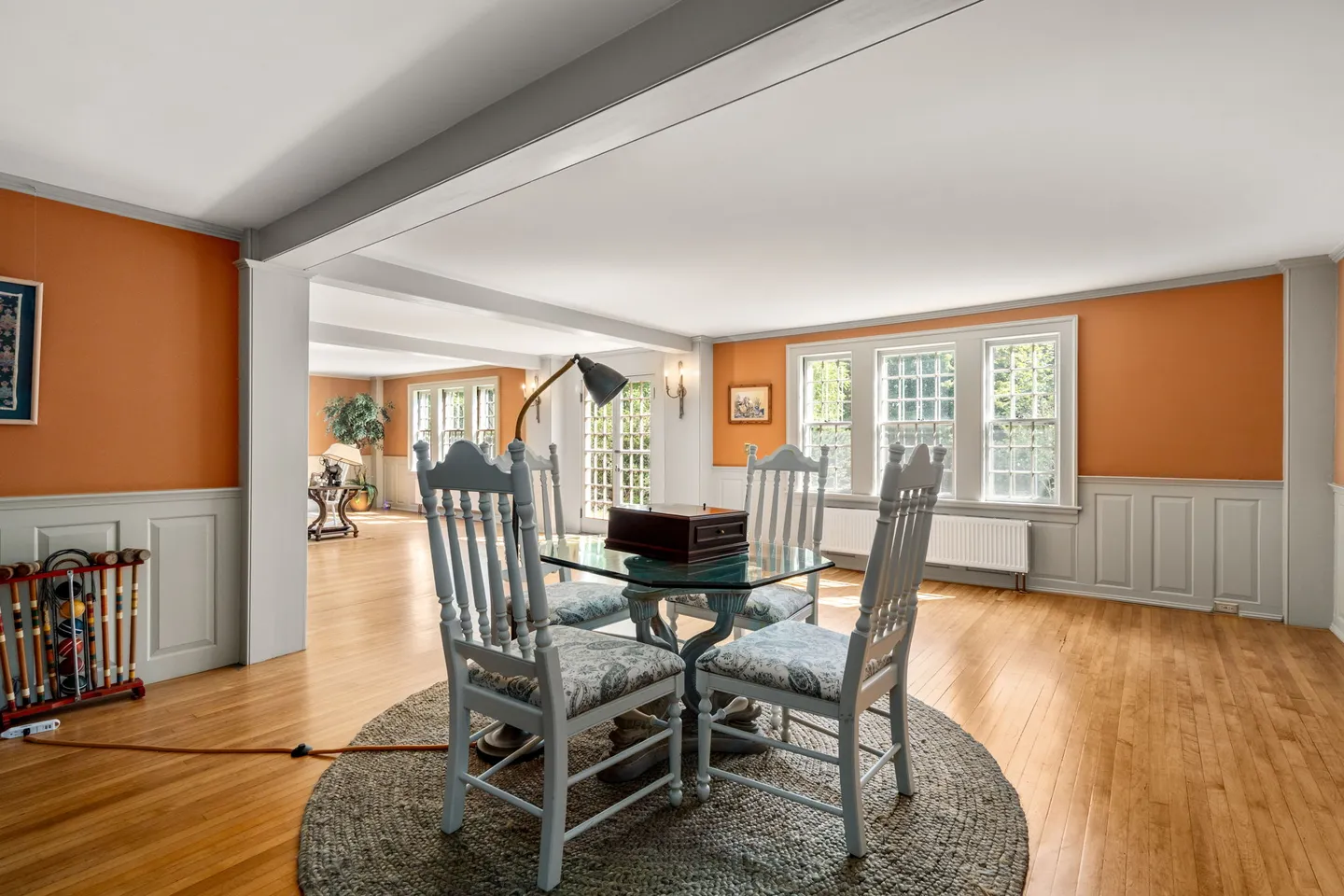 Dining area with terracotta-colored walls, light gray wainscoting, and a glass-topped table set on a circular rug over wood floors.