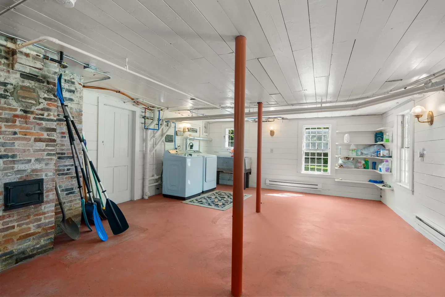 Basement utility and laundry room with painted plank ceiling, salmon-colored floor, exposed brick column, and washer-dryer set.