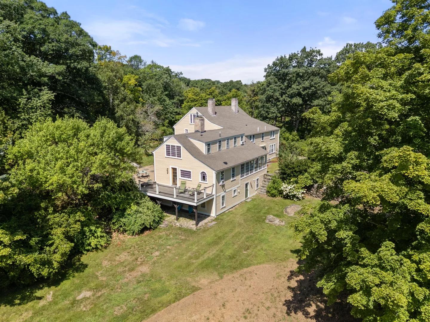 Elevated view of a large yellow multi-story house with a wooden deck and brick chimneys surrounded by dense green trees and a sloping lawn.