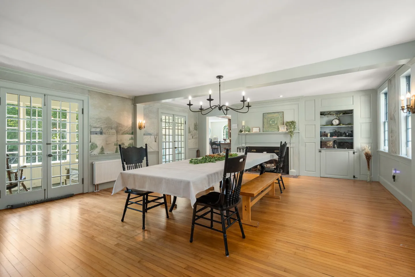 Spacious dining room with wood flooring, a long covered table with black chairs and a wooden bench, wall mural, and French doors.