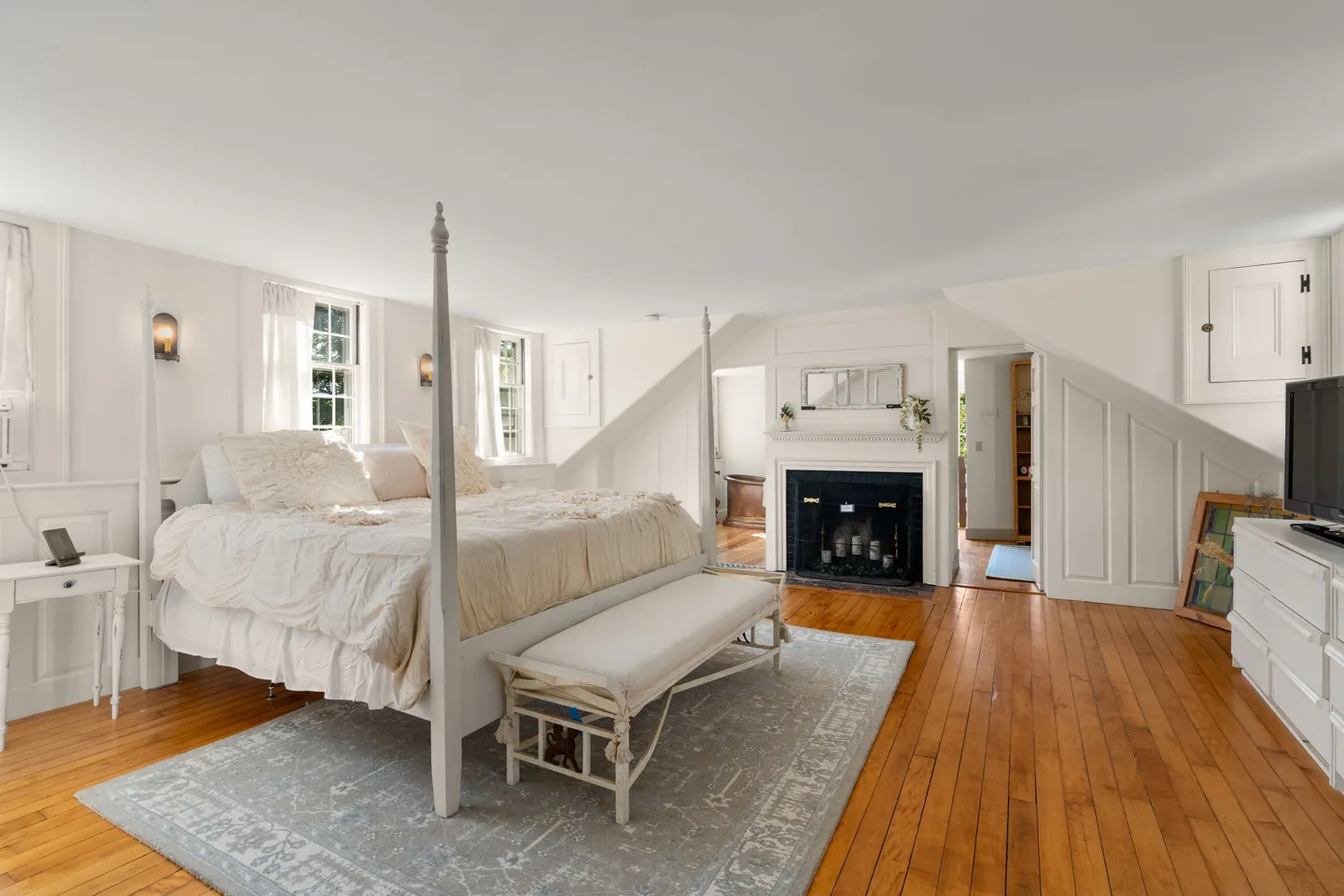 Spacious bedroom with wood floors, a four-poster bed, an upholstered bench, and a non-working fireplace with candles.