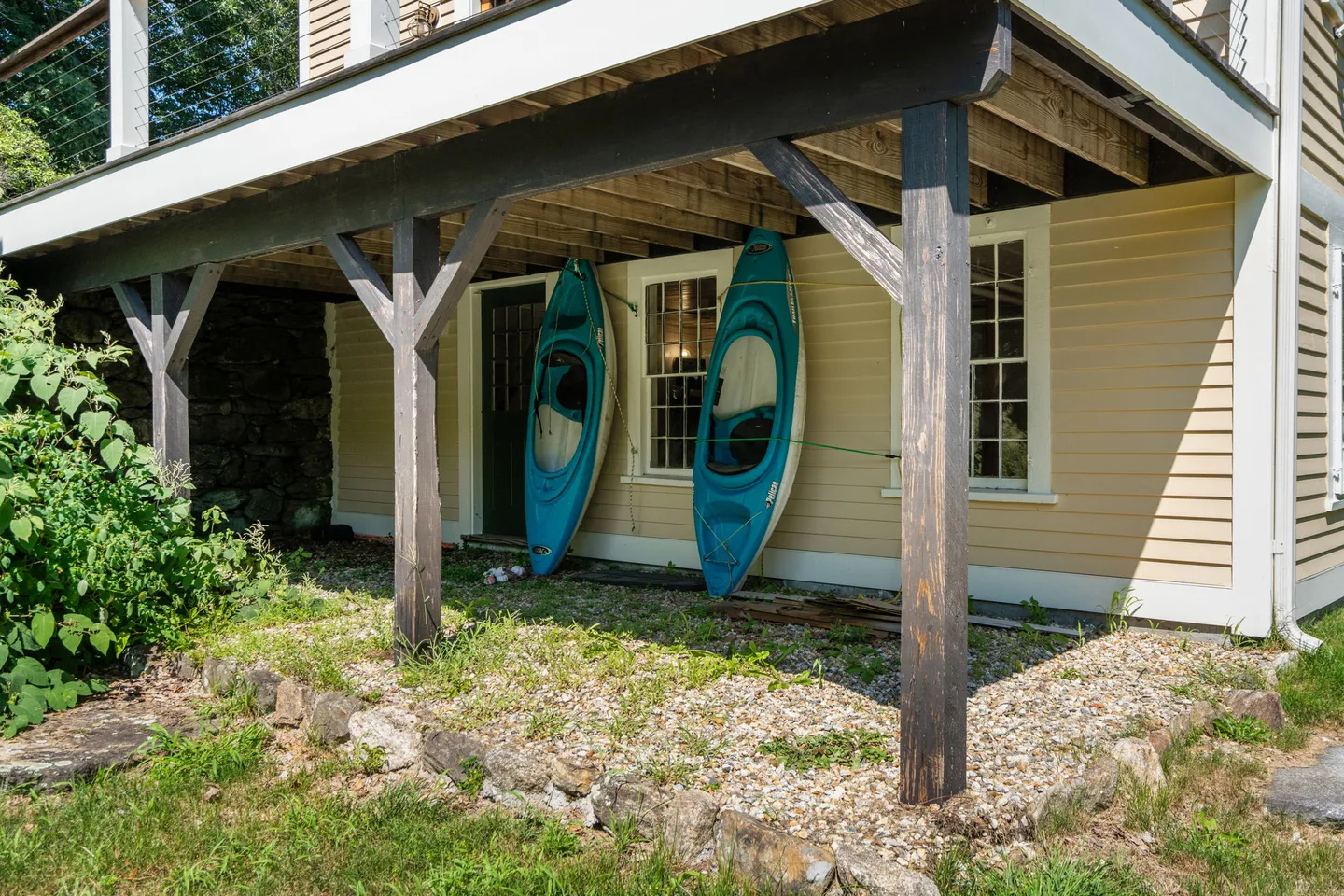 Two blue kayaks stored upright underneath a wooden deck support next to the siding of a yellow house.