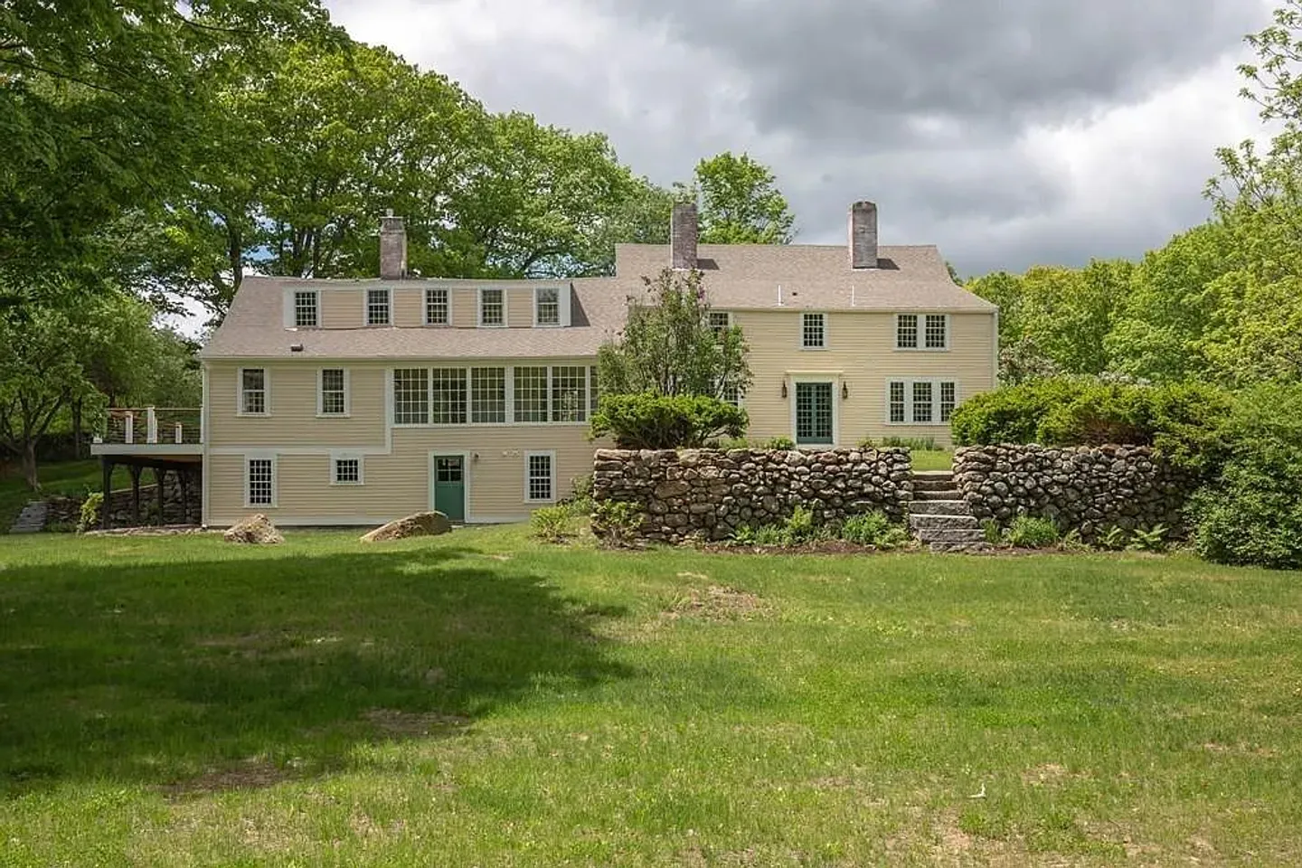 Exterior rear view of a two-story light yellow house with green trim, a multi-windowed sunroom, stone retaining wall, and green lawn.