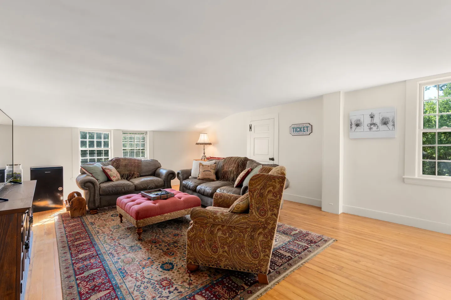Living room with sloped ceiling, leather sofas, patterned armchair, pink tufted ottoman, and wood flooring.