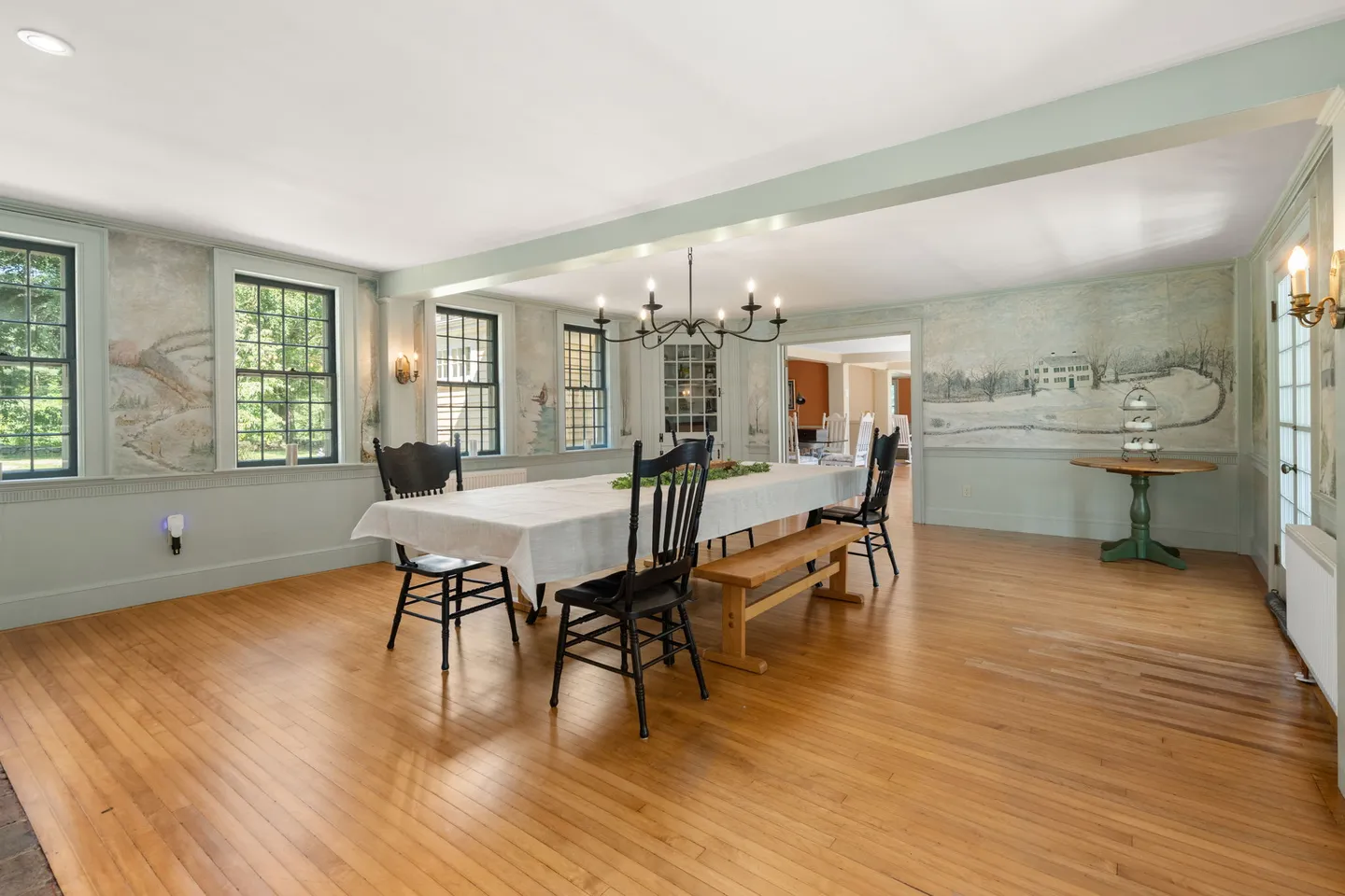 Spacious dining room featuring hardwood floors, landscape murals on the walls, and a long dining table set beneath a black chandelier.