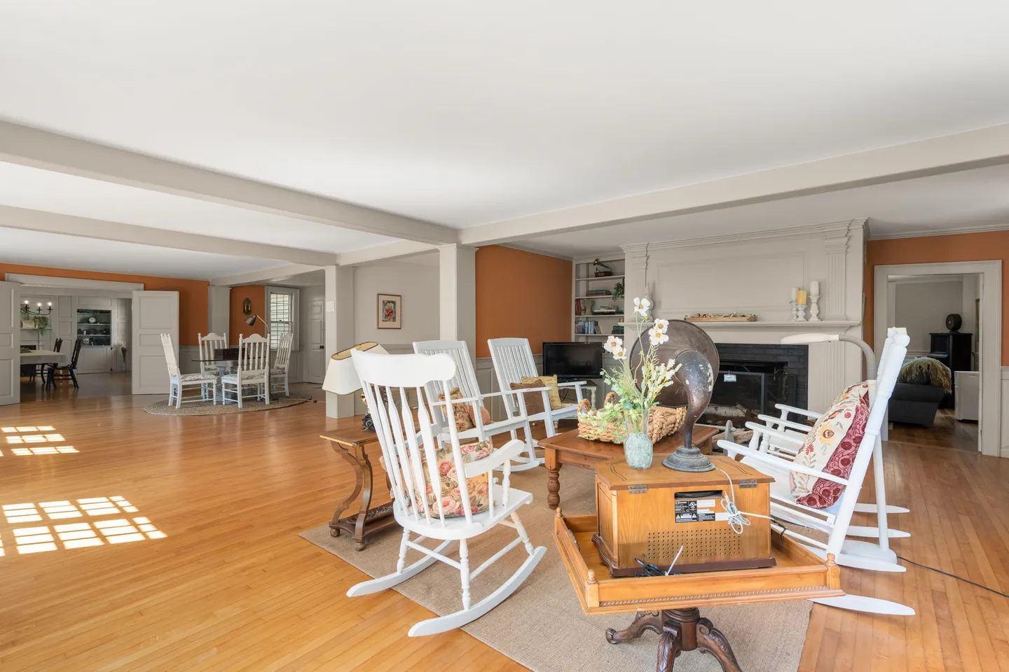 Spacious living room with light wood floors, a brick fireplace, and white rocking chairs arranged on an area rug, opening to a dining area in the background.