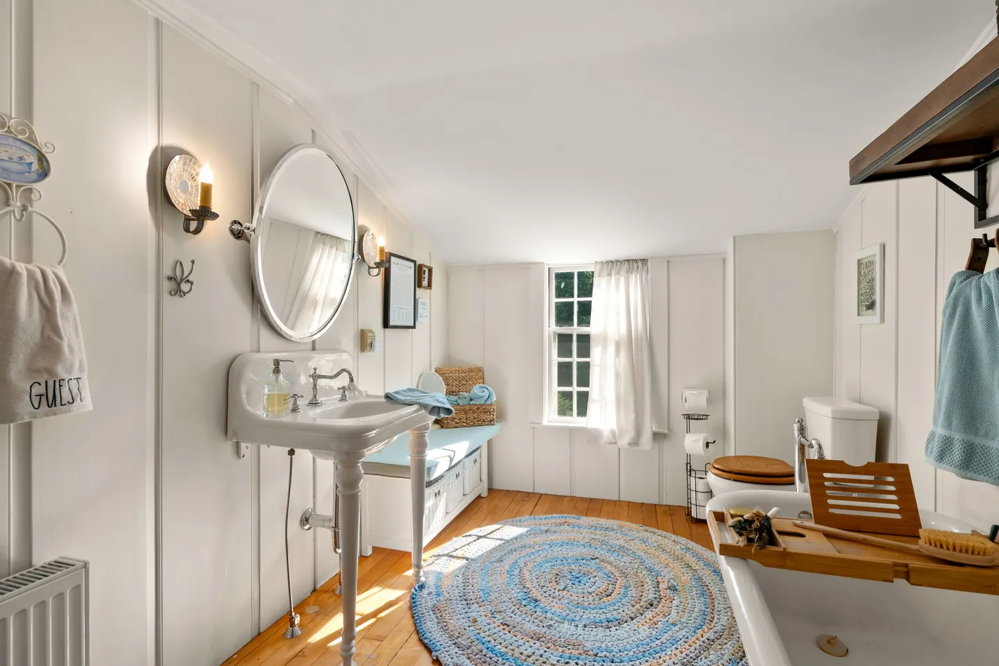 Bathroom with white wood-paneled walls, wood floors, a vintage pedestal sink, an oval mirror, a bathtub with a tray, and a window.