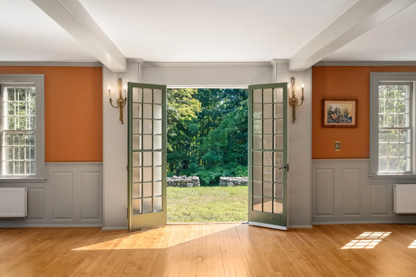 Open French doors leading from a room with orange walls, wainscoting, and wood flooring to a grassy yard with trees.