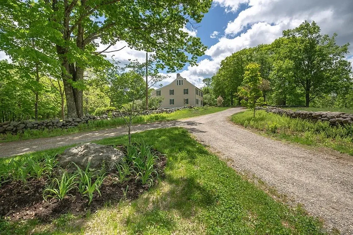 A gravel driveway curves past stone walls, green lawns, and mature trees toward a light-colored multi-story house under a blue, partly cloudy sky.
