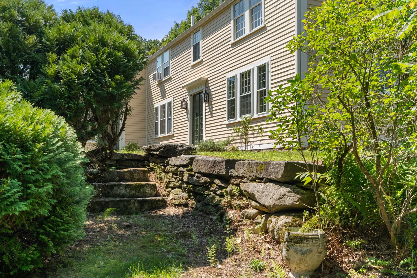 Exterior view of a beige-sided house with stone steps leading up a retaining wall past lush green foliage.