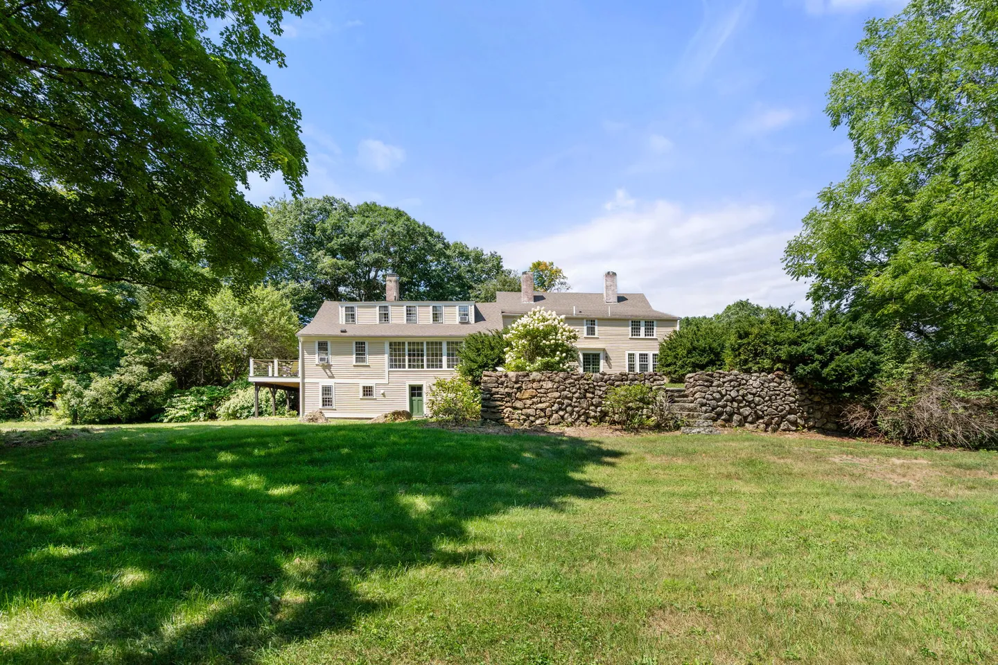 Exterior view of a multi-story home with light siding, surrounded by a large green lawn, mature trees, and a stone wall.
