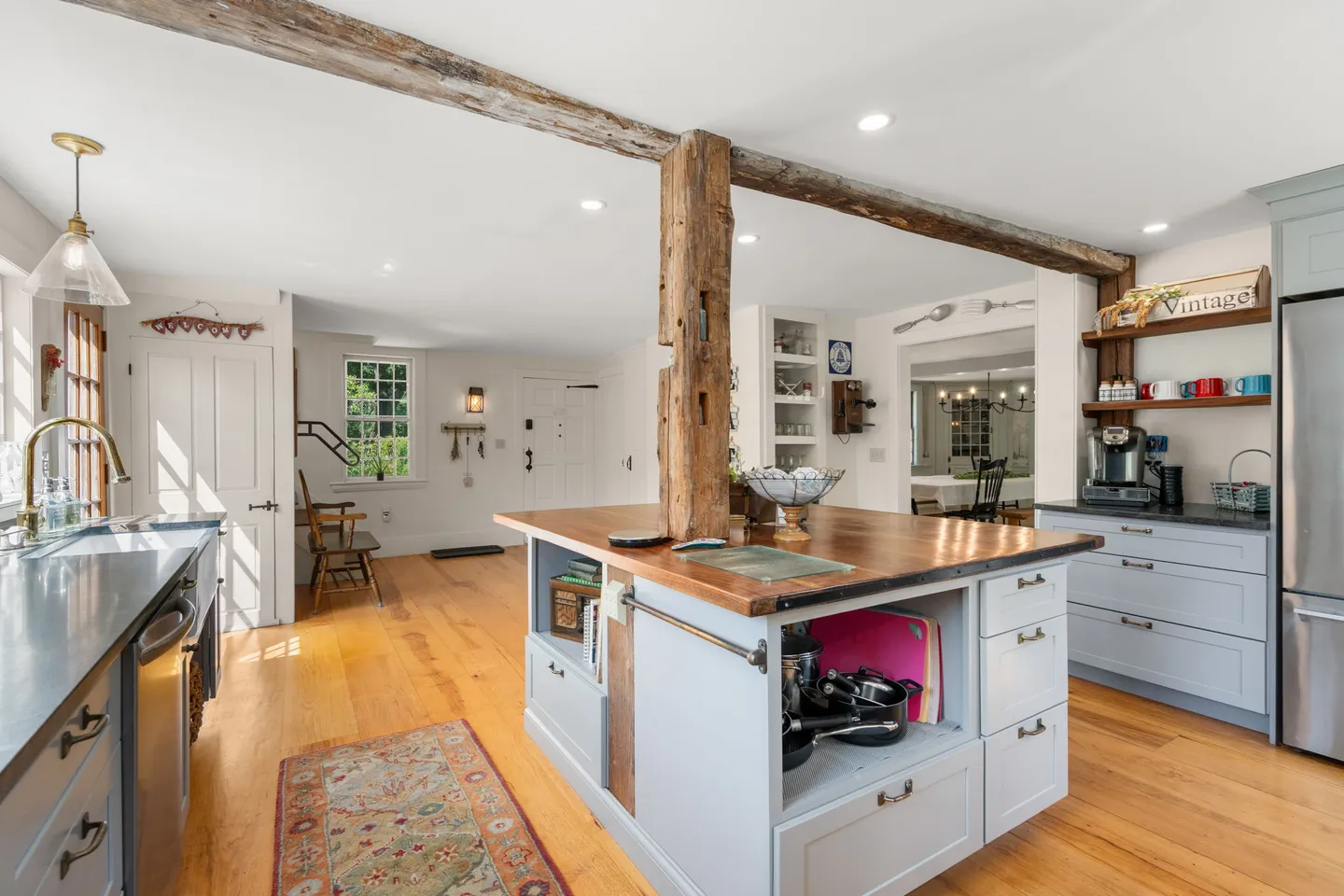 Bright kitchen featuring a central island with a wood countertop built around an exposed rustic timber support beam, light gray cabinetry, and wood floors.