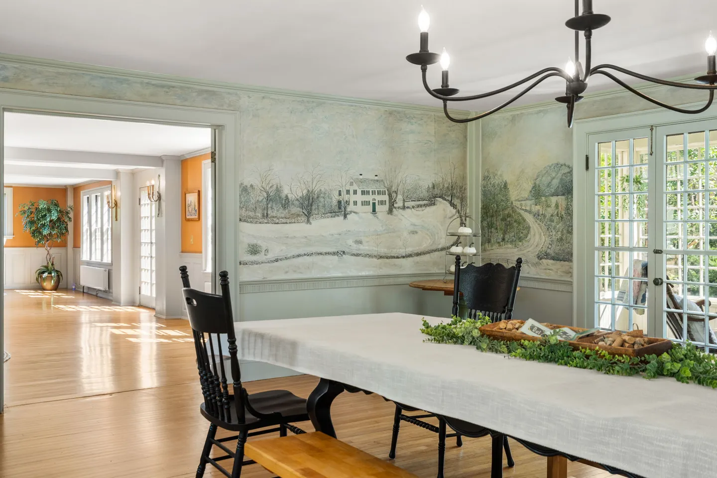 Dining room with painted scenic wallpaper murals, a long dining table, wooden bench, chandelier, and a wide doorway into an adjacent hallway.