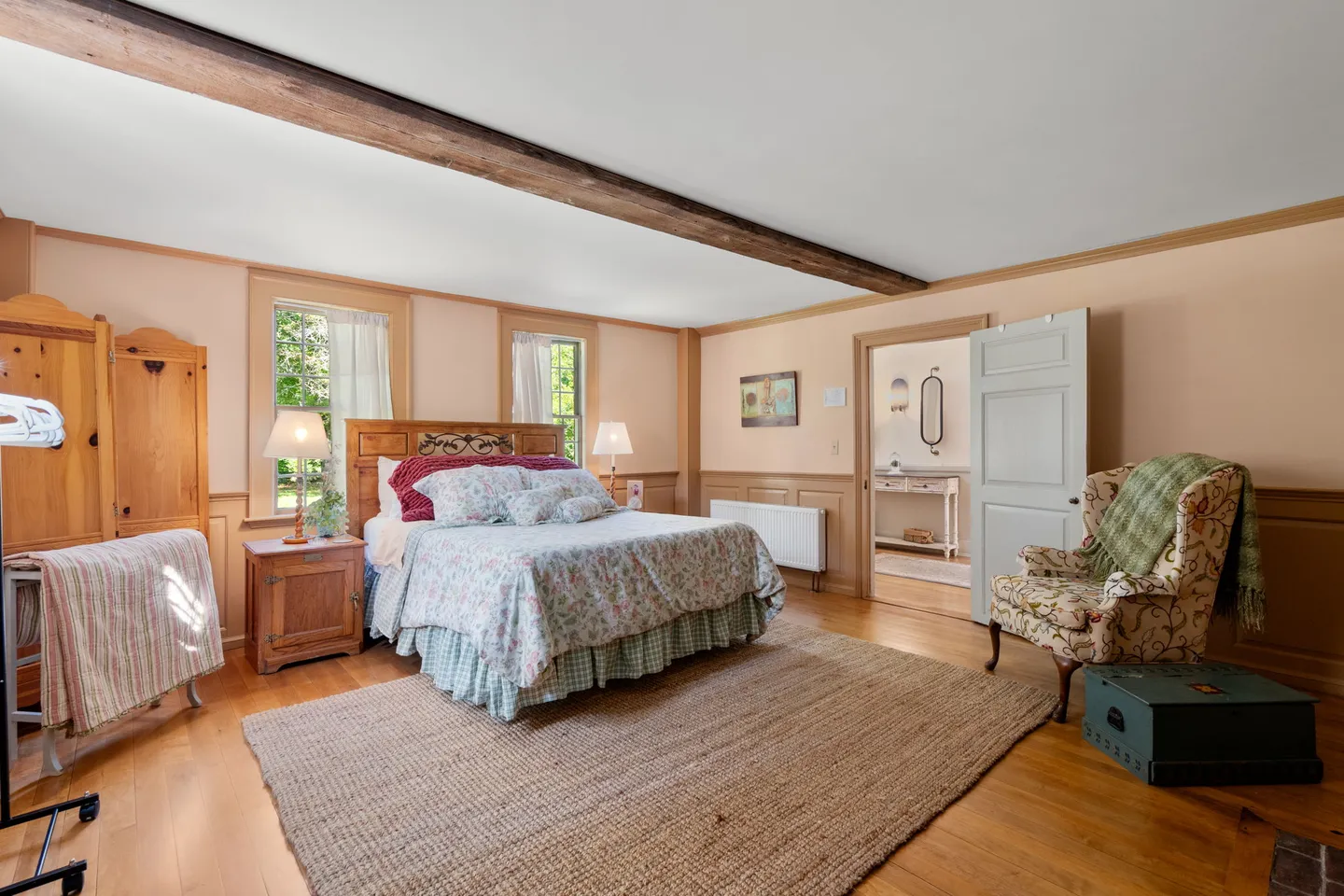 Bedroom with hardwood floors, an exposed ceiling beam, a wooden bed, patterned armchair, and an open doorway leading to a hallway.
