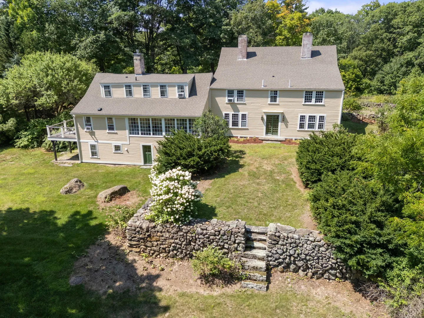 Elevated view of a light-yellow multi-story colonial home with a grassy lawn, stone retaining wall with steps, and surrounding green trees.