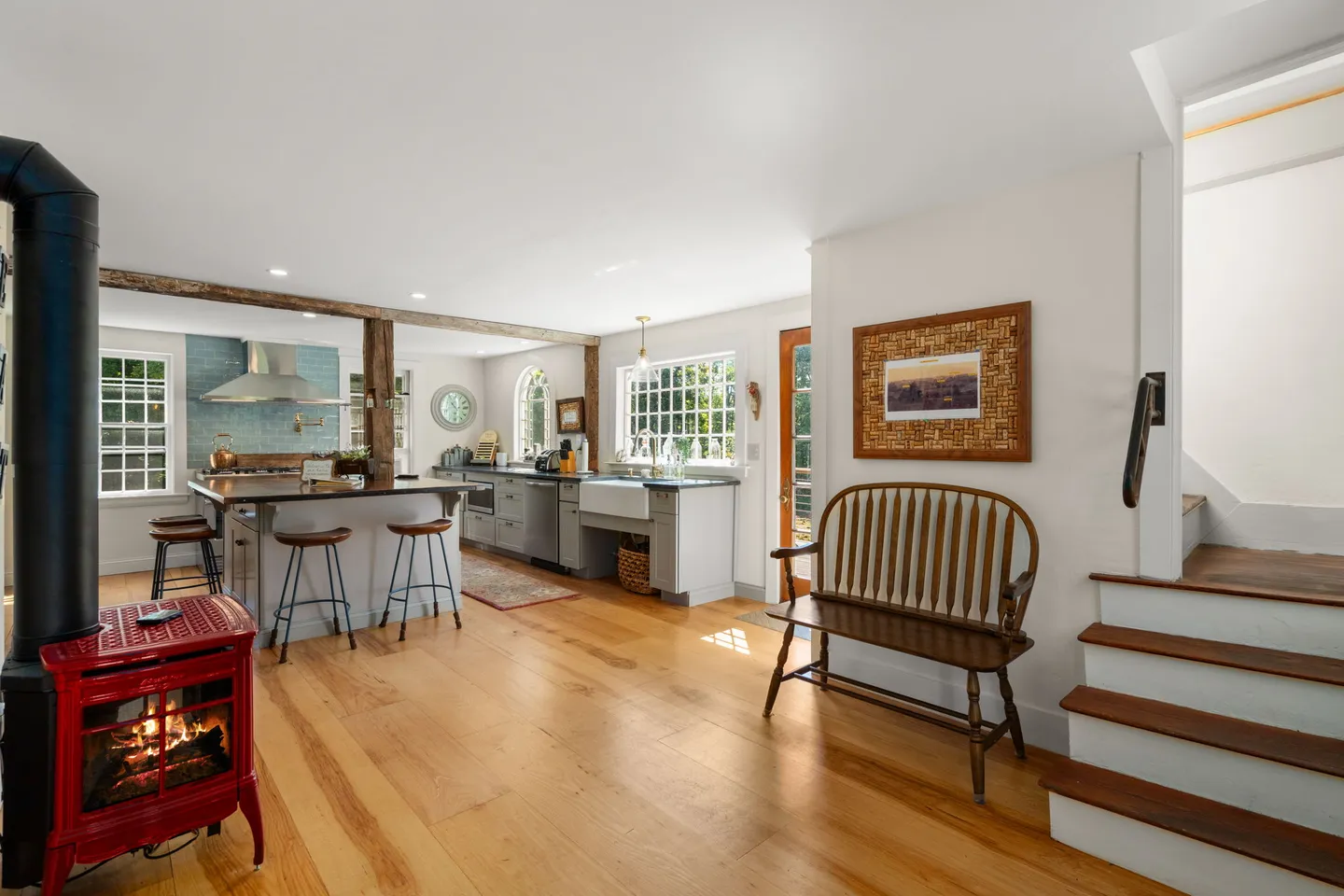 Open-concept living space and kitchen with light wood floors, a red freestanding stove, wooden bench, and staircase.