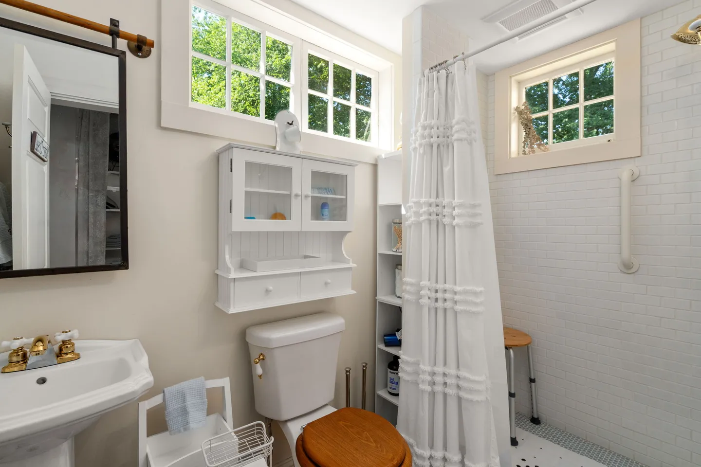 Bathroom with a pedestal sink, wall-mounted mirror, toilet, white storage cabinet, and a walk-in shower with white tile and a curtain.