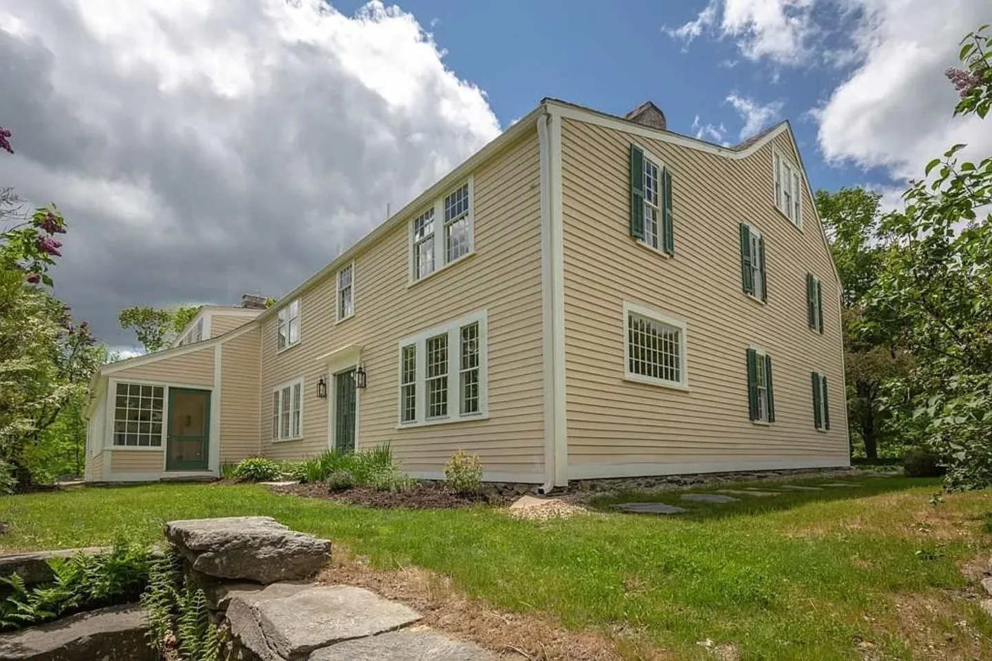 Exterior of a two-story light yellow house with green shutters, white trim, and a grassy lawn under a partly cloudy sky.