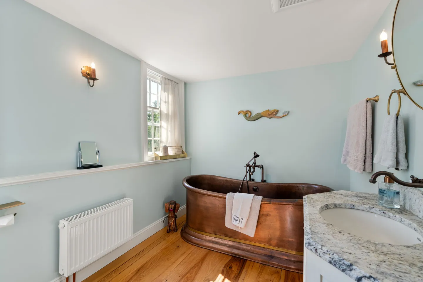 Bathroom with light blue walls, a copper soaking tub, a vanity with a stone countertop, and light wood flooring.