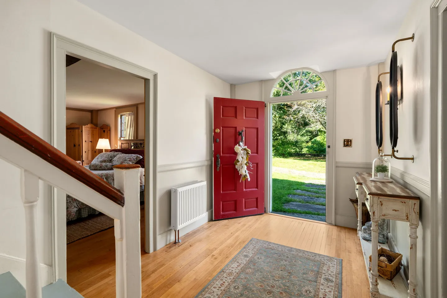 Entryway with an open red front door topped with a fanlight window, hardwood flooring, and a view into an adjacent bedroom.