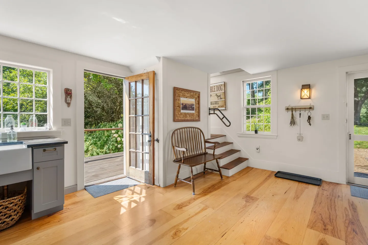 Sunlit entryway and mudroom area with light wood flooring, white walls, a wooden bench, and open glass-paned doors leading outdoors.