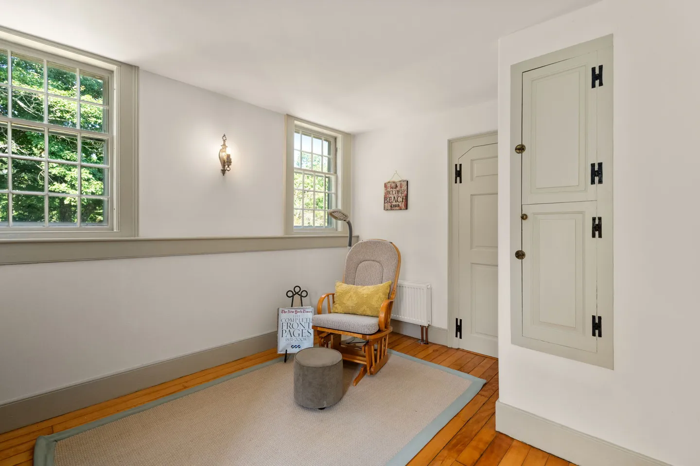 Seating area with wood flooring, an area rug, a rocking chair with an ottoman, and two windows.