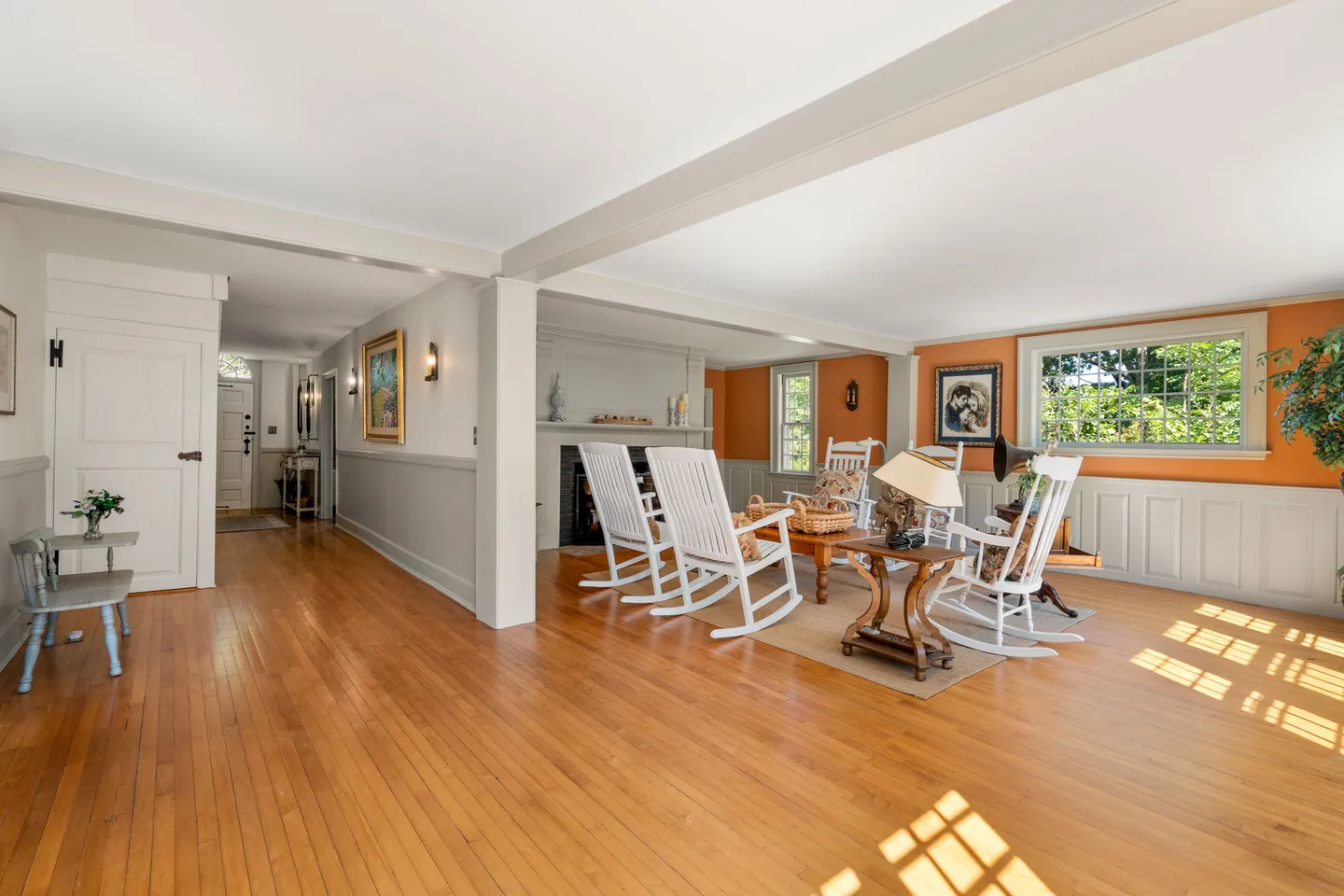 Spacious living room with wood flooring, white rocking chairs around a low table, a fireplace, and an orange accent wall with wainscoting.