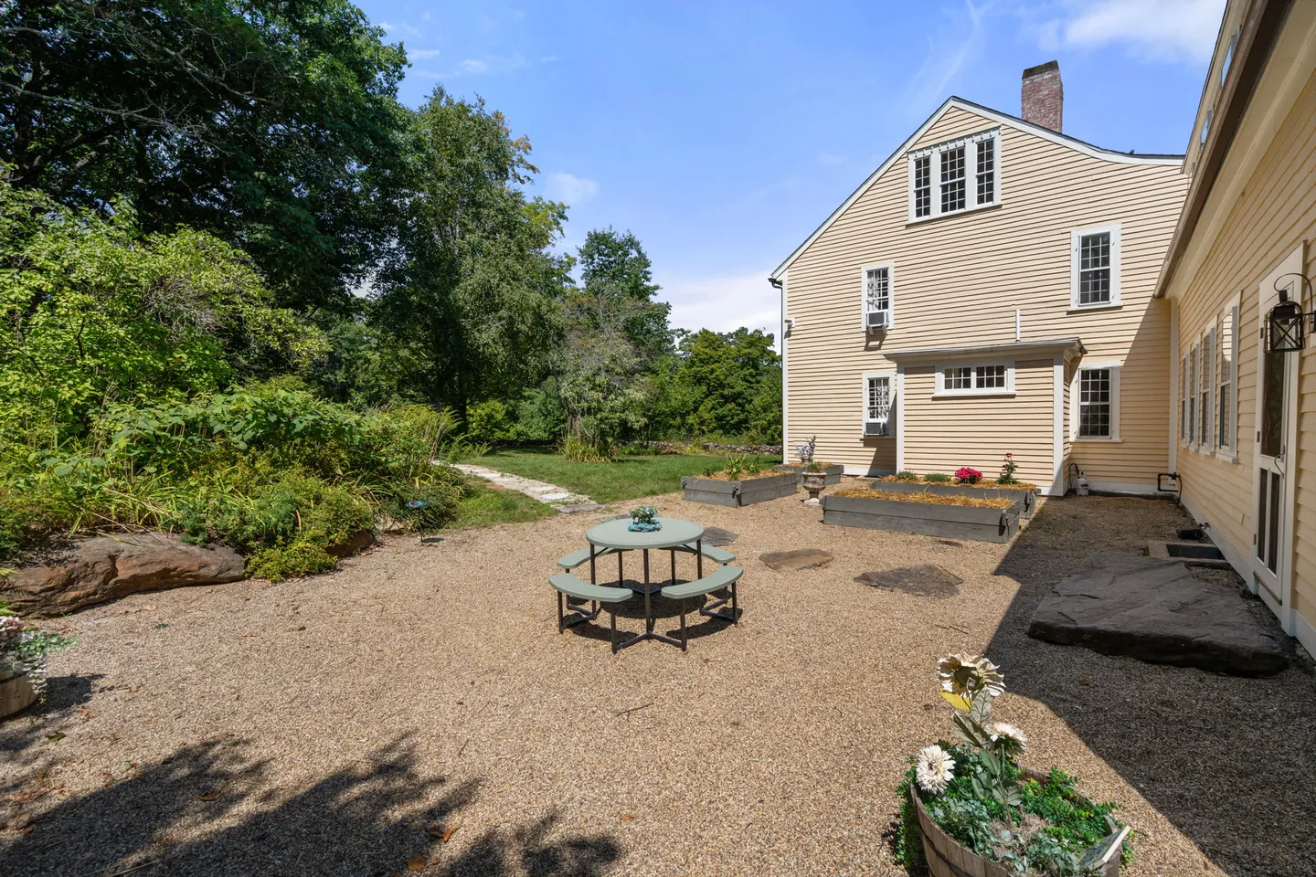 A gravel patio with a circular picnic table beside a yellow siding house with a green lawn and trees in the background.