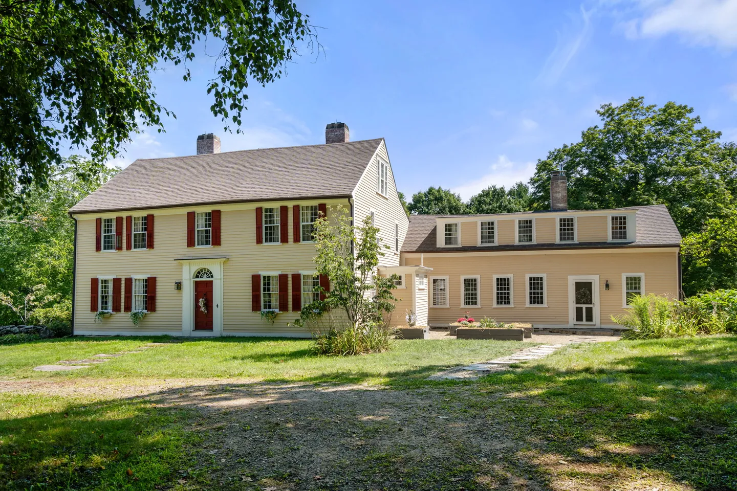 Exterior front view of a two-story yellow colonial-style house with red shutters and an attached side wing on a grassy lawn.