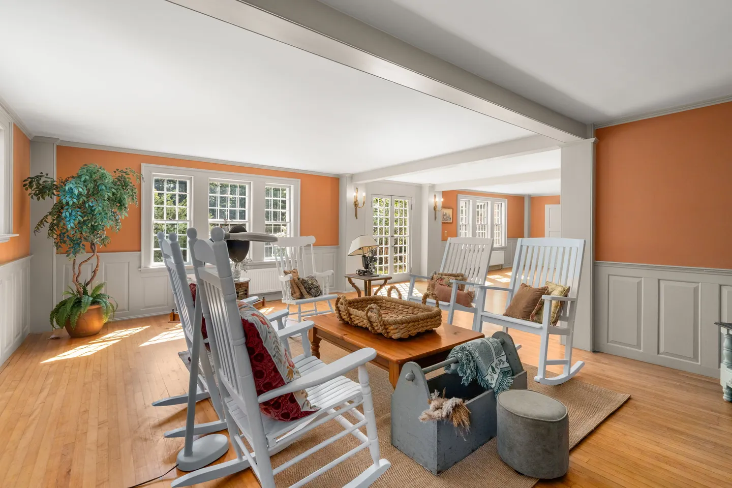 Sunlit living room with terracotta-colored walls, grey wainscoting, wood flooring, and white rocking chairs arranged around a wooden coffee table.