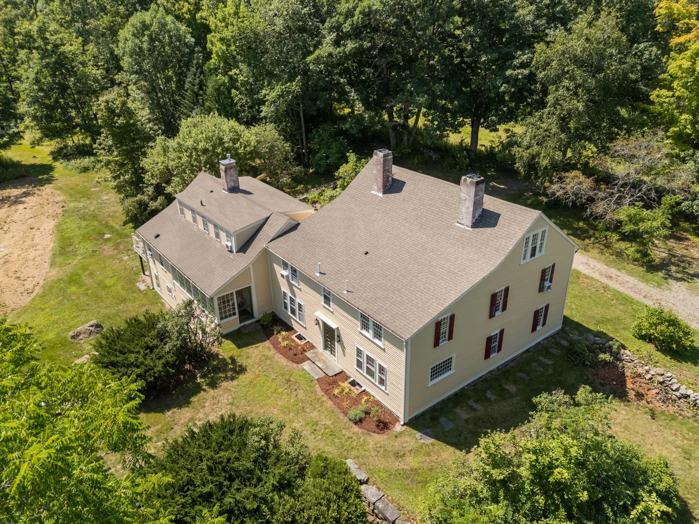 Aerial view of a two-story yellow farmhouse with brown shingled roofs and chimneys surrounded by lush green trees and a yard.
