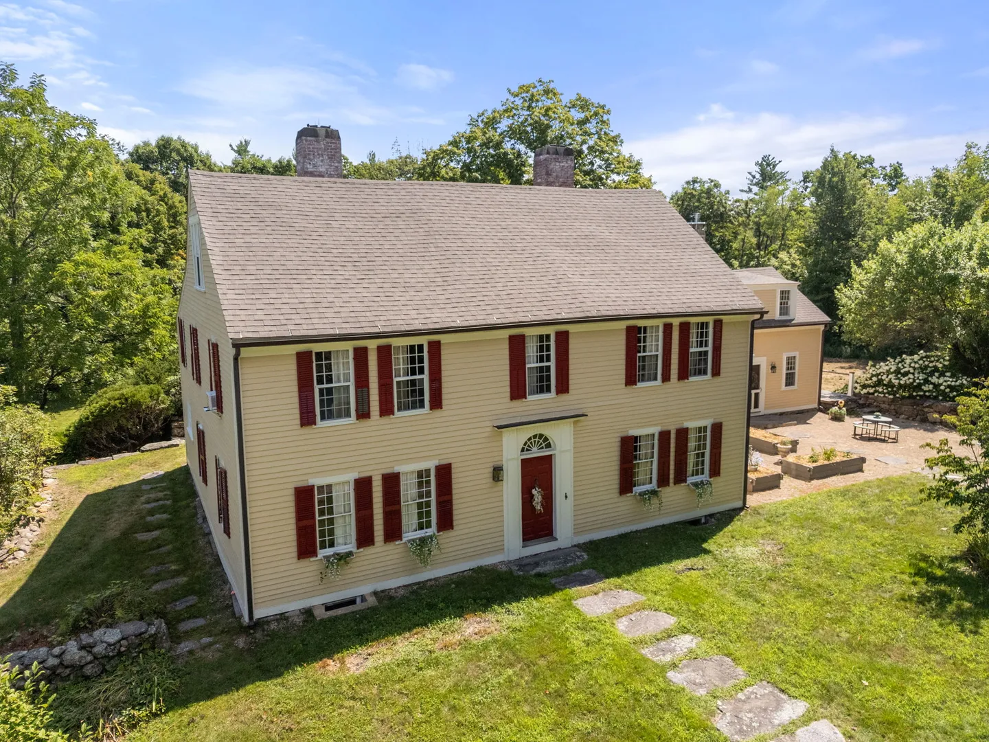 Aerial view of a two-story yellow house with a gabled roof, red shutters, and a stone walkway across the lawn.