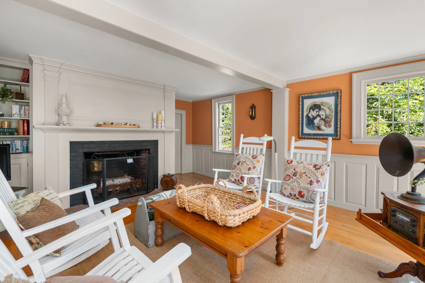 Living room featuring a black brick fireplace with a white mantel, white rocking chairs, a wooden coffee table, and orange accent walls with wainscoting.