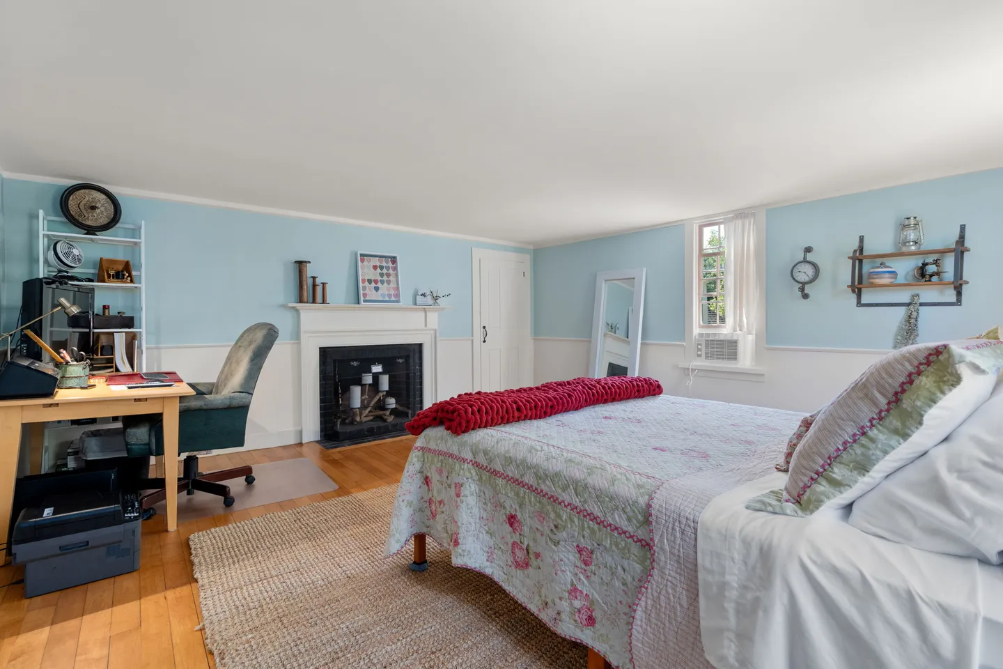 Bedroom with blue walls, a fireplace, wood flooring, a bed with a floral quilt, and a desk workspace.