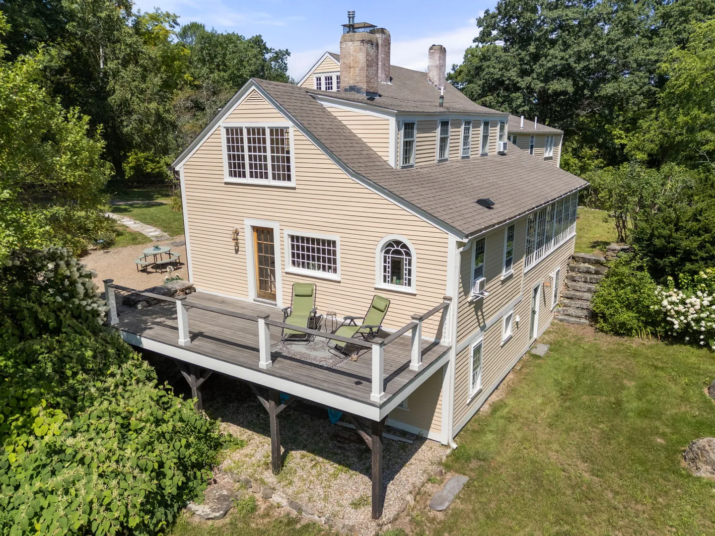Elevated view of a multi-story yellow-sided home featuring a raised wooden deck with lounge chairs surrounded by lush green trees.