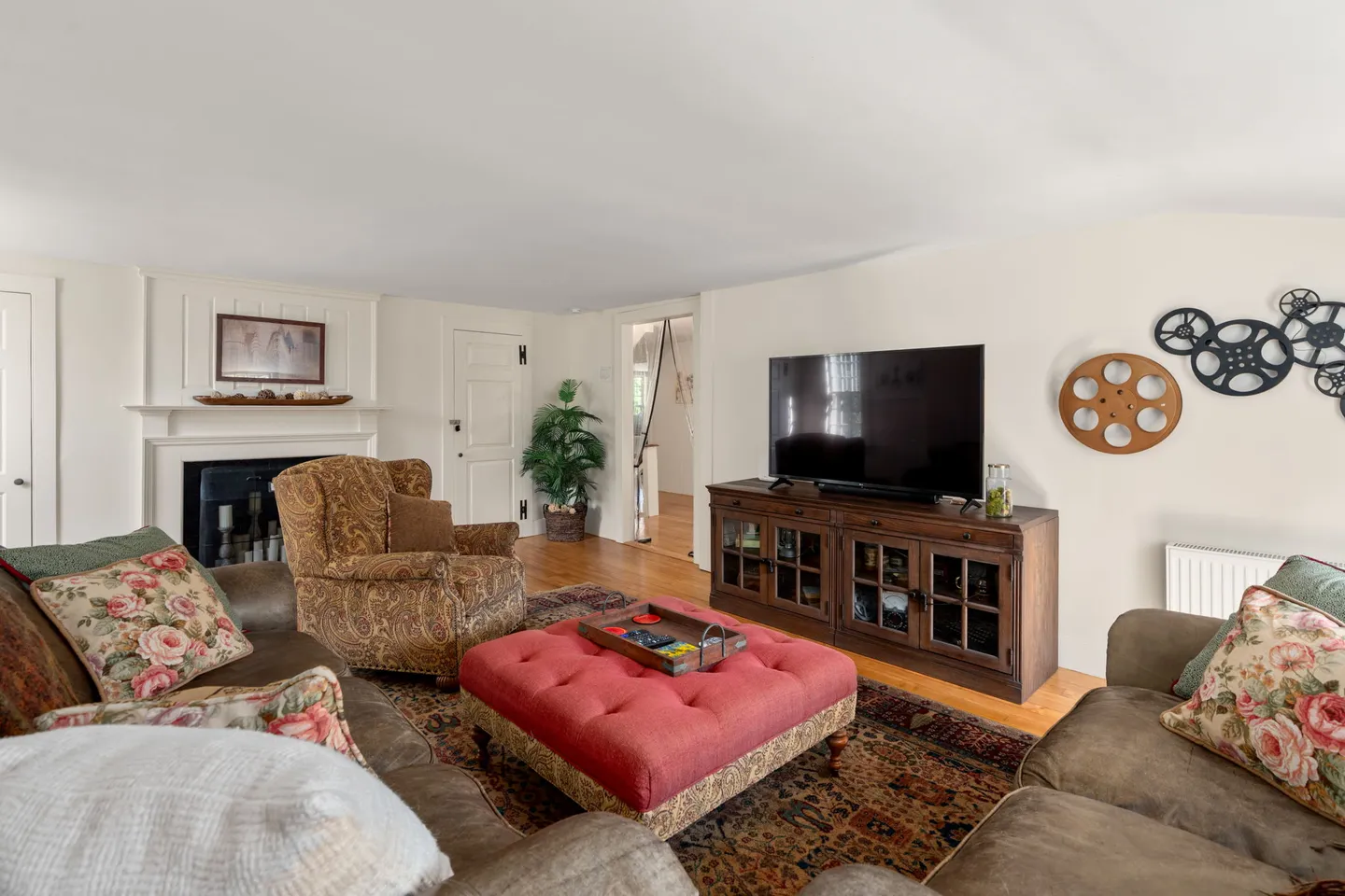 Living room with brown sofas, a floral armchair, a red tufted ottoman, and a flat-screen television on a wooden media console.